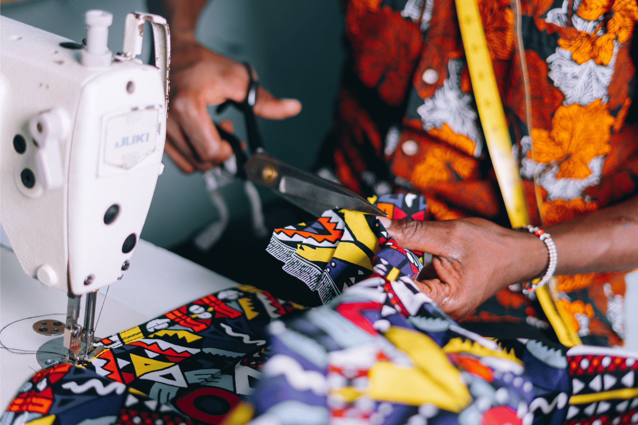 Person cutting fabric with scissors next to a sewing machine, wearing a colorful garment.