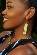 Model wearing gold rectangular earrings with a blurred green background