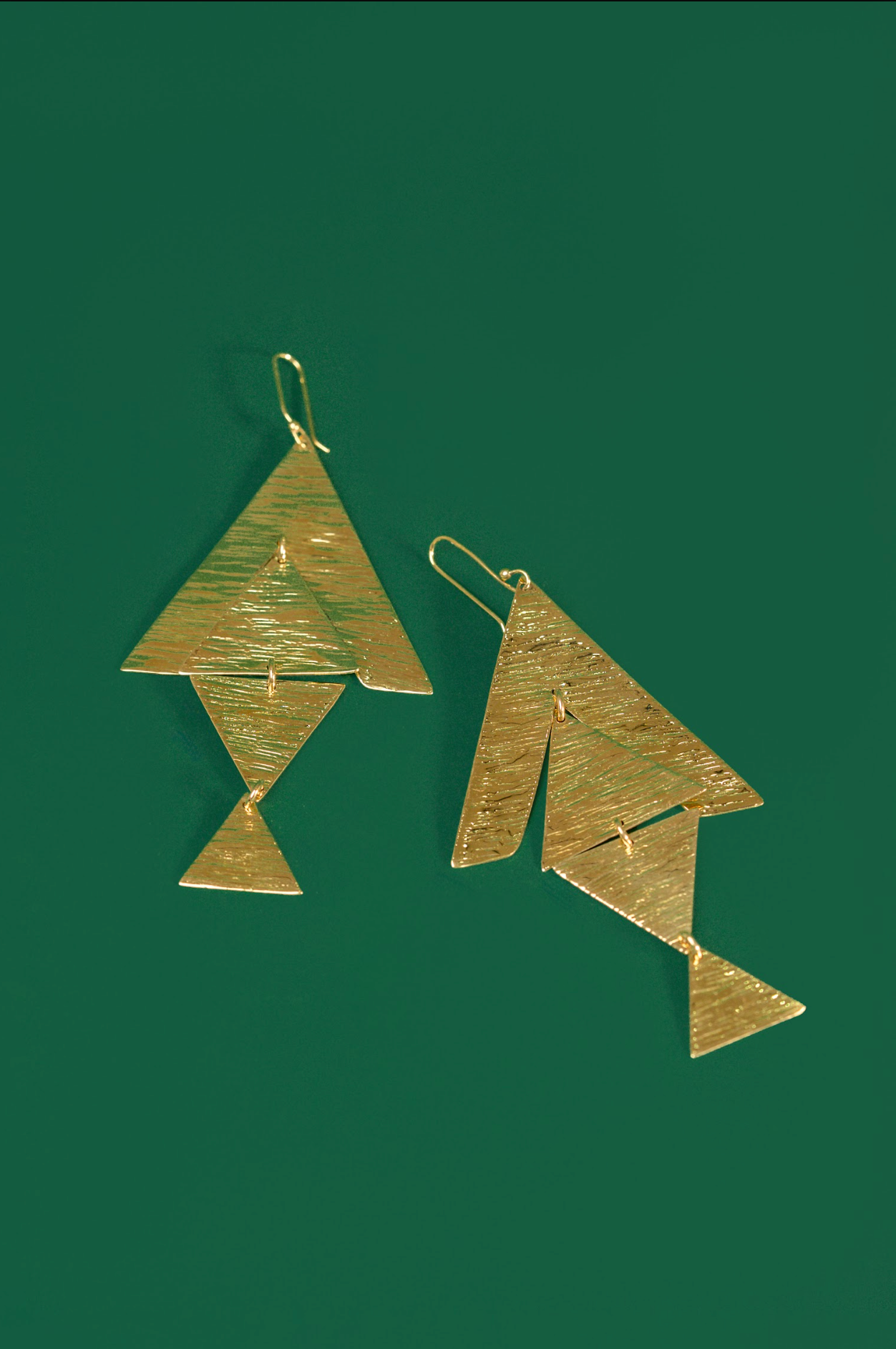 A pair of gold geometric dangle earrings with textured, hammered surfaces, displayed against a solid dark green background.